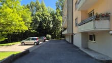 Multi-story apartment building with balconies, surrounded by trees and greenery, with a paved parking area in the foreground.
