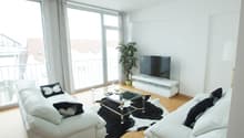 Spacious living room with large windows, white walls, hardwood floors, white leather sofas, glass coffee table, and a potted plant. The room has access to a balcony or terrace.
