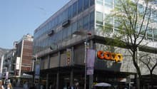 Modern building with glass facade, large sign reading 'COOP', multiple floors, people walking in front