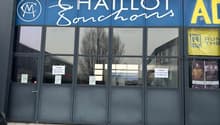 Commercial building, gray walls, large glass doors, parking area in front, signage in French