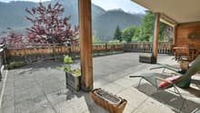Wooden balcony or terrace with a scenic view of the mountains and forests. The balcony has a concrete floor, wooden railings, and some potted plants. There are chairs and other outdoor furniture on the balcony, suggesting it is a relaxing outdoor living s