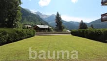 The image shows a scenic mountain landscape with a large grassy lawn in the foreground. There are several wooden chalets or cabins visible, with balconies or terraces. The mountains in the background are covered in lush green forests. The sky is blue with
