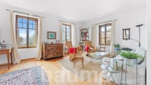 Living room with white walls, wooden floor, beige rug, large window, wooden cabinet, couch, books on shelves, coffee table, lamp