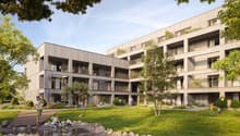 Multi-story apartment building with wooden exterior, balconies, and surrounded by trees and greenery. There is a grassy area with a small pond or water feature in the foreground, where children are playing.