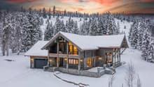 Log cabin-style house with a sloped roof, stone foundations, multiple windows, a driveway, balcony, and snow-covered surroundings