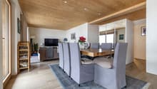 Dining room with wooden table, gray chairs, wooden ceiling, carpet, living room with couch, TV, plants, and a mirror.