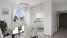 Open-concept living area with dining table and chairs, kitchen with white cabinets and appliances, large windows with curtains providing a view of the outdoors, and a modern light fixture above the dining table.