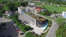 A large multi-story commercial or office building with a curved glass facade, surrounded by a parking lot and green landscaping. The building appears to be situated in a rural or suburban area with a view of a small town or village in the background.