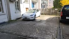 The image shows a white Tesla Model 3 electric vehicle parked on a cobblestone driveway in front of a multi-story residential building. The building has a balcony and there are trees visible in the background, indicating an urban setting.