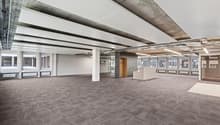 Large open office space with exposed concrete ceiling and beams, gray carpeted flooring, and several windows along the walls. The space appears to be a commercial or office environment, with a clean and modern design.