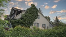 Two-story house with a gabled roof, surrounded by lush greenery and vegetation. The house has shuttered windows and a balcony or terrace visible on the upper level. The exterior walls are painted a light color, and the roof appears to be made of tiles or
