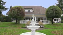 Large house with brown roof, white walls, large windows, white porch, garden with fountain, trees, and grass