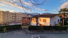 a single family house surrounded by hedges and greenery, a big building in the background, mountains in the distance