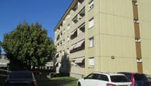 A multi-story building with beige walls and brown trims. It has awnings on the balconies. There is a tree and a play area nearby. There are 3 cars parked in front of the building.