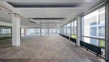 Empty office space with large windows, grey carpet flooring, white ceiling, wooden floor, radiators, support columns.