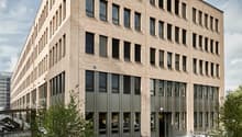 Multi-story office building with a modern, rectangular design featuring large windows and a paved outdoor seating area with tables and chairs.