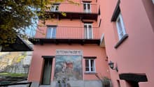 A two-story pink building with balconies, surrounded by trees. The building has a mural painting on the front and a set of stairs leading to the entrance.