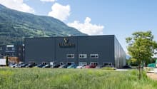 Large black industrial-style building with the Valmont logo, surrounded by a grassy area and parked cars in the foreground. The building appears to be a commercial or industrial facility located in a mountainous, forested area.