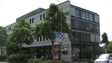 Multi-story office building, white exterior, glass windows, ground-level storefronts, trees and bushes in front, cars parked on street, street light in front.