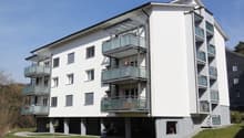 A white building with multiple floors, featuring several balconies, raised ground floor, and bicycles parked on the ground floor.