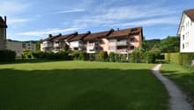 A row of apartment buildings with brown roofs and balconies, set against a backdrop of greenery and a wide grassy field.