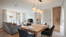modern dining area with wooden table and chairs, gray sofa, table set for 6, large windows, tv, wooden cabinets