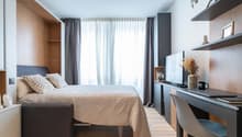 Large bed, decorative pillows, white blanket, wooden walls, white curtains, desk, monitor, cabinet, chair, shelves with frames, flower arrangement, and mug