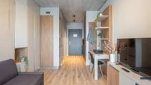 Open-plan studio apartment with concrete ceiling, wooden floors, built-in storage, desk area, sofa, and a balcony or terrace visible.