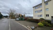 Apartment building, concrete path, shrubs, bushes, trash bins, streetlight, cars driving