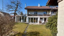 2-story house, brown roof, large glass windows, balcony, garden, potted plants, pathway, grassy lawn, trees, nearby houses