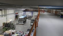 The image shows a large industrial facility or warehouse. The space is filled with various equipment, machinery, and storage racks. There are several workstations and production lines visible. The ceiling is high and the lighting is industrial. The overal
