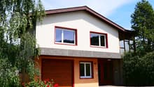 2-story house with white exterior walls, red-framed windows, and a wooden garage door. The house has a balcony or terrace on the upper level. The front yard features a paved driveway and red flowers in the landscaping.
