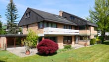 2-story house with a slanted roof, brick and wood exterior, large windows, balconies, and a well-maintained garden with a red Japanese maple tree in the front yard. There appears to be a parking area or driveway in front of the house.