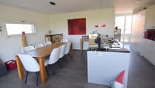 Dining room with a long wooden table, modern chairs, gray tiles, open kitchen with countertop, and sliding doors to the balcony.