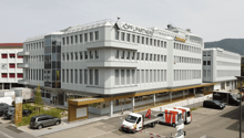 multi-story office building, white exterior, glass windows, red sign reading 'KOPFLI PARTNERS', multiple parked vehicles in the front