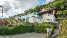 House on slope, glass balcony, terrace, brick ground floor, driveway, front and side gardens