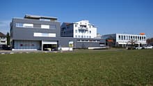 Commercial building with sign 'Migros', grey exterior, multiple windows, open garage door, cars parked in front, spacious lawn area