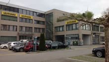 Commercial building with multiple signs reading 'LEUTHHARD', several cars parked in front, two trash bins, building has large windows, and paved surface