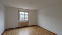 An empty room with white walls, wooden floor, a window, and a radiator.
