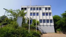A white, three-story office building with a ground floor, multiple windows, and a street-facing side. There is an entrance with blue railings and a sign on the left side. A pole is standing in front of the building, and a tree is growing next to it. In th