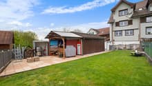 Two-story residential building with a tiled roof, surrounded by a grassy yard and a paved patio area. The yard features a wooden shed or storage structure, a barbecue grill, and other outdoor furniture. There is also a garage or carport visible on the pro