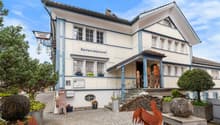 Two-story building with a traditional German architectural style, featuring a restaurant called 'Speiserestaurant' on the ground floor. The building has a tiled roof, large windows, and decorative elements. There are some rustic metal sculptures of animal