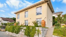 Two-story house, yellow exterior, white windows, stone wall, large staircase, fenced area, green plants, orange vase.