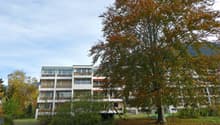 Multi-story apartment building with balconies, surrounded by trees with autumn foliage, grassy area in the foreground