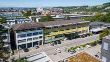 Large office building with yellow and green exteriors, numerous windows, parking lot with cars parked, solar panels on the roof, surrounded by greenery and other buildings
