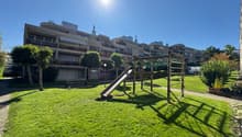 Playground with a slide and a bench, surrounded by green grass and trees, with a walkway leading to the building