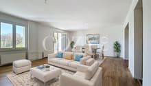 Spacious open-plan living room with large windows, hardwood flooring, beige sofa set, coffee table, and dining area with table and chairs. The room has a bright and airy feel with natural light and potted plants.
