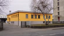 A single story yellow building with gray trims. A gate leads to the parking lot with trees in the center. Several windows and a chimney are visible on the yellow wall. A grey building is on the right side.