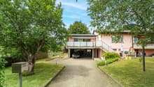Two-story house with a pink exterior, surrounded by lush greenery and trees. The house has a balcony on the upper level and a garage with a parking space underneath. The property has a paved walkway leading to the entrance and a well-maintained lawn.
