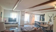 Living room with wooden floor, large sofa, dining table, TV, kitchen area, wooden beams on ceiling
